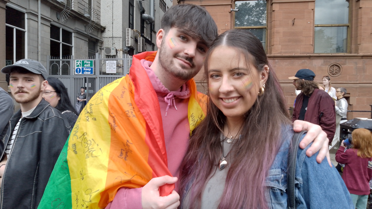 Robyn and Max standing next to each other smiling at the camera. Max has is arm around Robyn and is draped in a pride flag