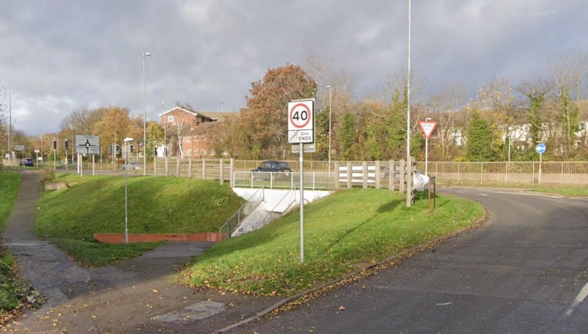 A road on the right curving round to the left, and entering a roundabout. There are railings either side of the carriageway on the roundabout. There is a white 40 Zone ENDS sign in the foreground and a Give Way sign beyond that. There is a sloping pavement on the left which enters a white walled underpass beneath the roundabout.