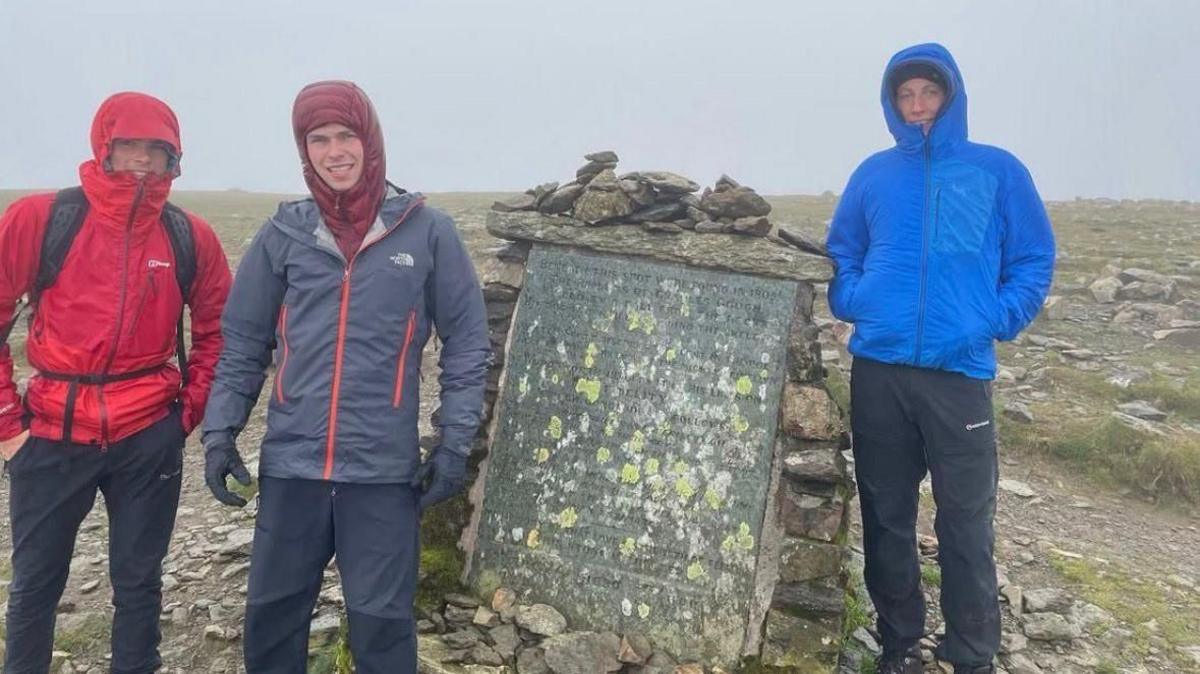 Mae tri dyn ifanc mewn siacedi gaeaf coch, llwyd a glas yn sefyll ar gopa mynydd wedi'i orchuddio â chymylau
