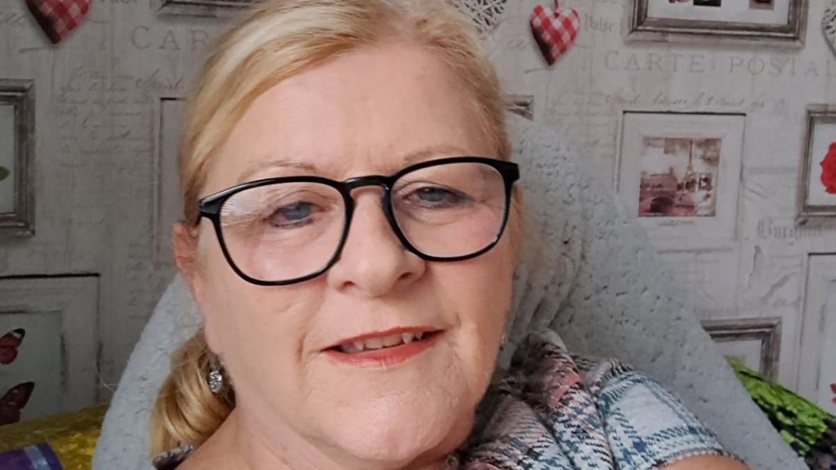 Wendy Swain at home. She has glasses on and is smiling.  She has blonde hair, pulled behind her head in a ponytail and has blue eyes. Her mouth is slightly open. Behind her on a wall decorated with wallpaper with grey writing, are a series of framed pictures.