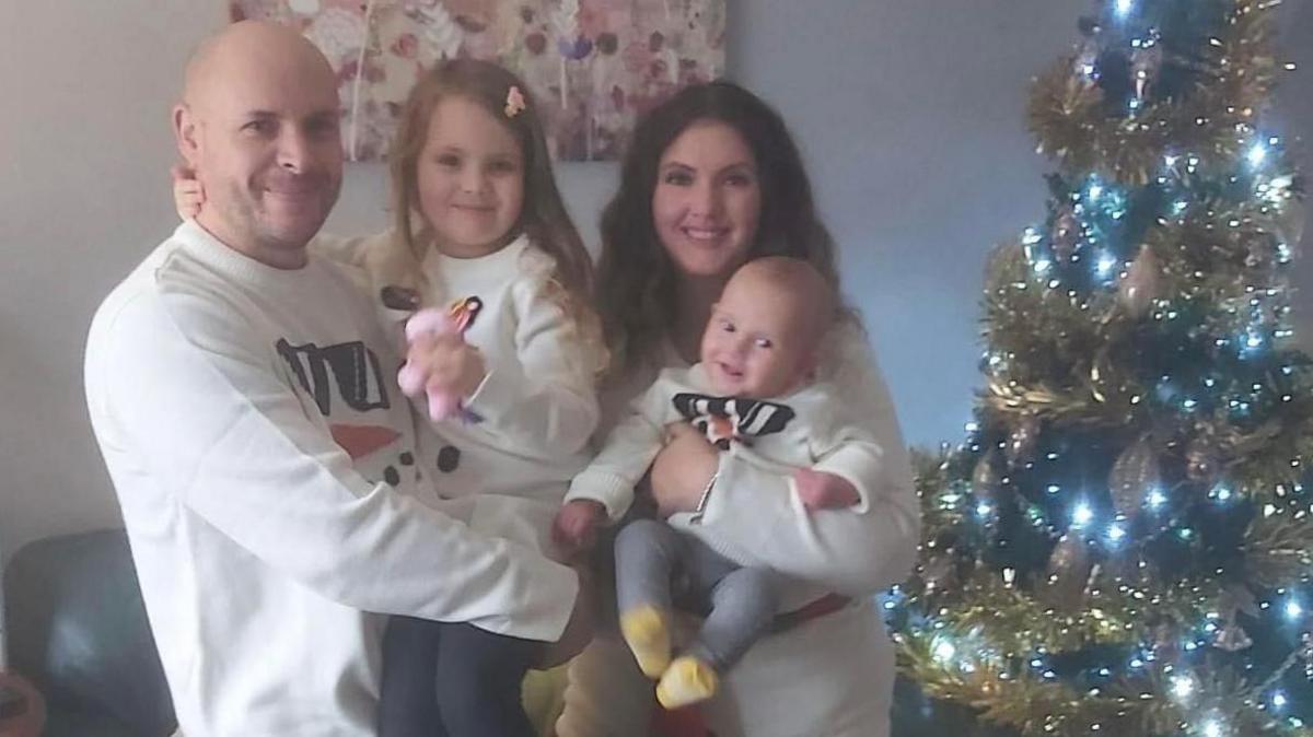 Christian and Naomi holding their two young children. One is a toddler and the other is a baby. They are standing next to a Christmas tree.