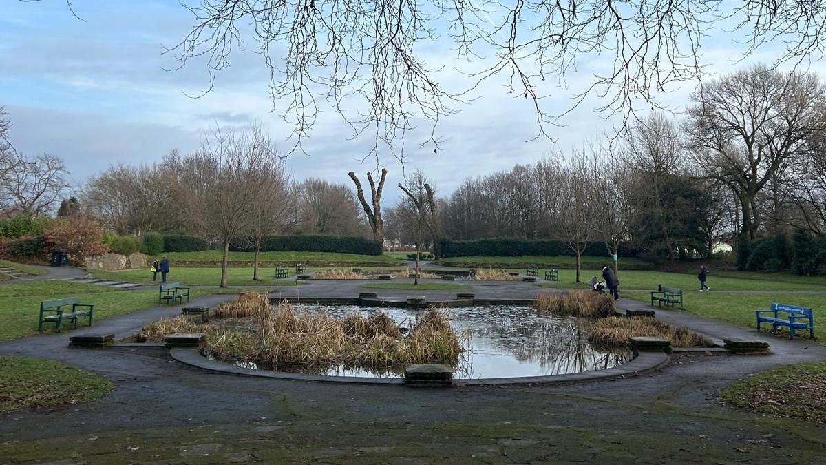 A large pond surrounding by a circular path, park benches and green fields. 