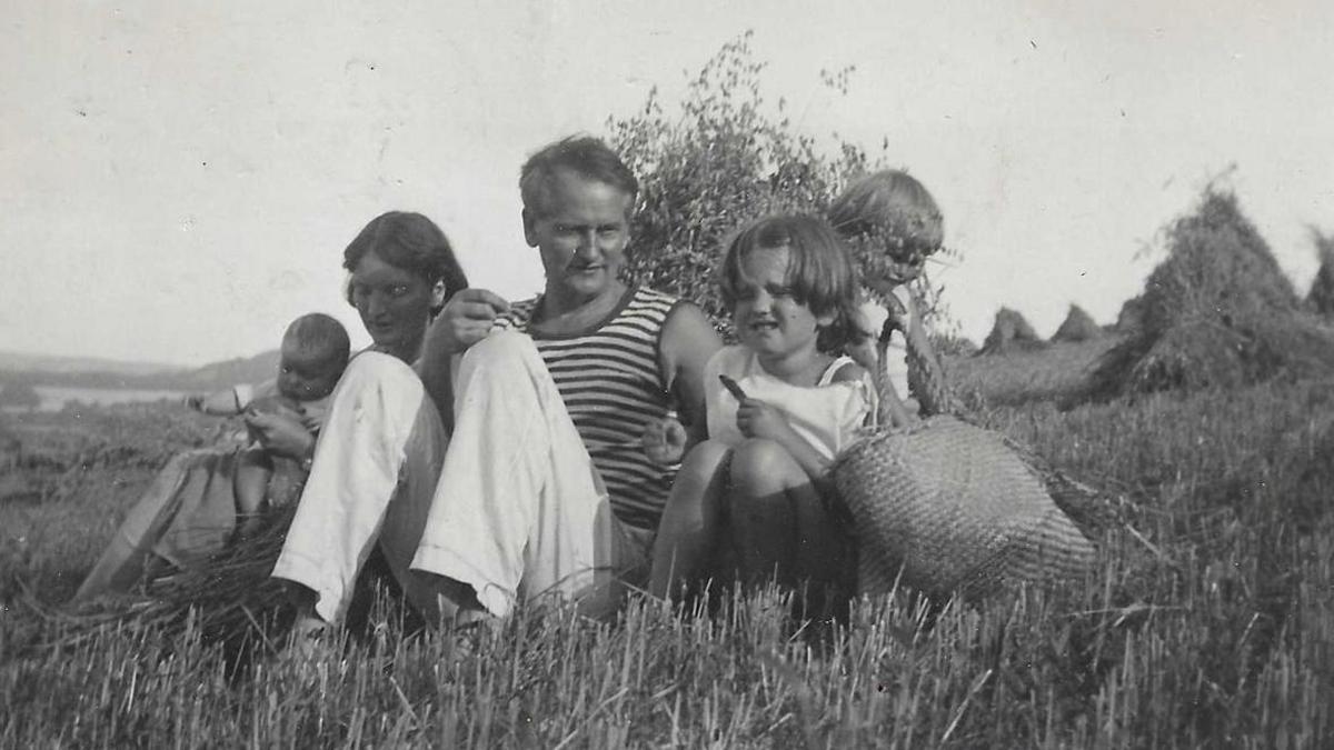 A black and white photo of Noel Carrington, with a woman and three children in a field. They are all sitting down with hay around them and a child holding a wicker bag.