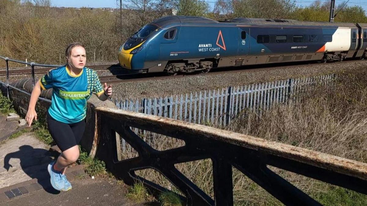 A woman wearing a blue running top which reads 'Samaritans' on it, black shorts and blue trainers, with her blonde hair tied back, runs across a bridge. A blue train is on a track underneath the bridge, which reads 'Avanti West Coast' on the side of it. It is a sunny day.
