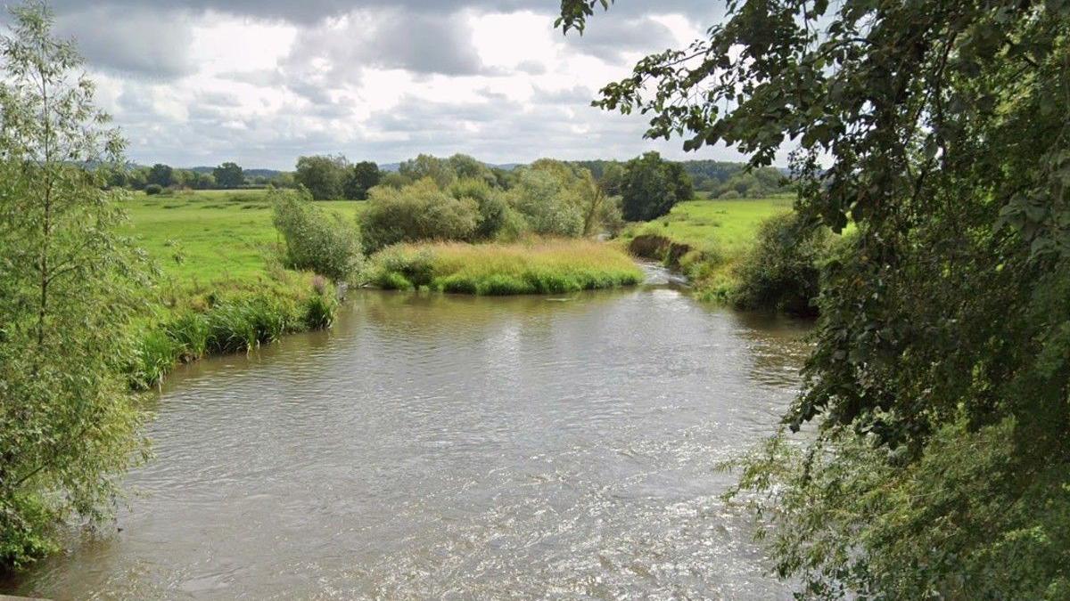 The River Lugg from Wergins Bridge near Sutton St Nicholas. The photograph shows a winding river flowing through countryside and fields and shrubs and trees overhanging the water.