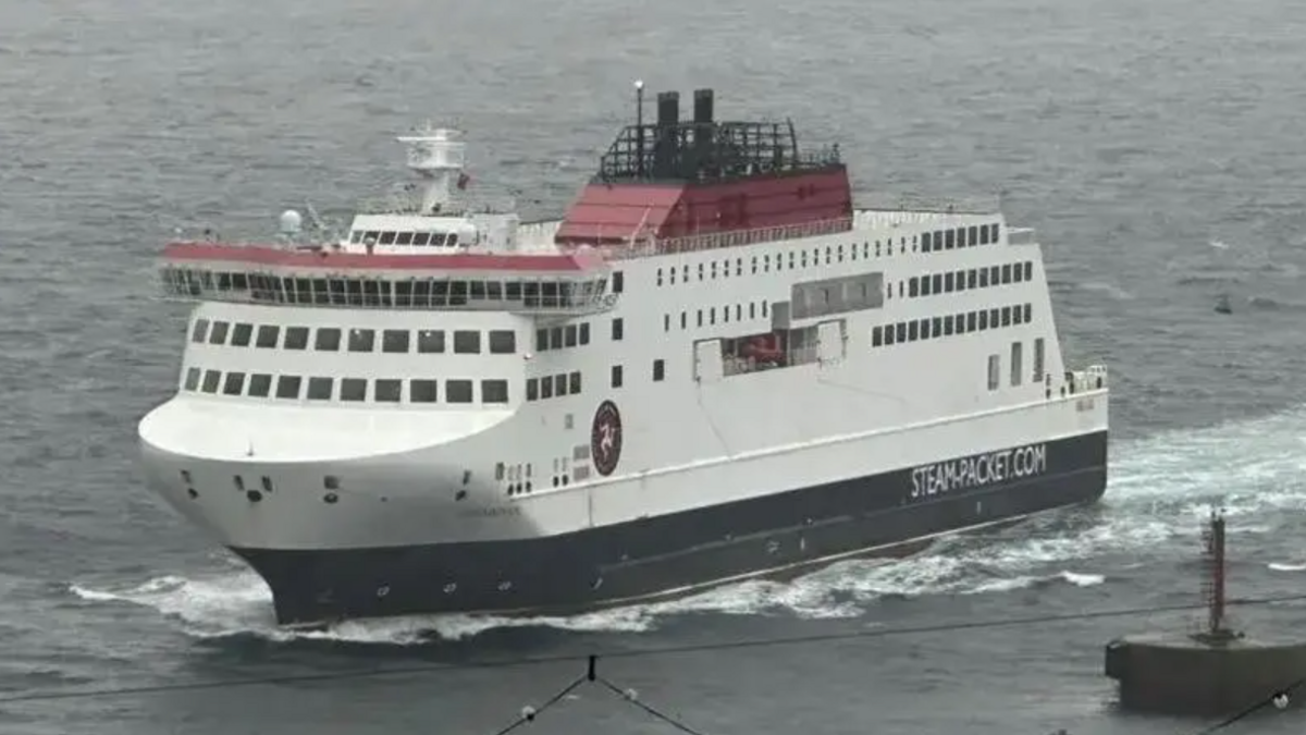 The Manxman ferry, a tall white ship, with red detail on the top and dark base, driving through waves at sea. A buoy can be seen on the right and some festoon street lighting in the close foreground on a grey day.