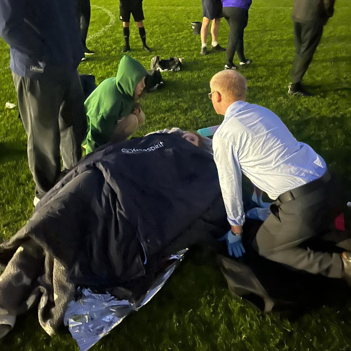 Two people kneel around a woman covered in a pile of jackets lying on the grass of a football pitch. It appears to be the evening