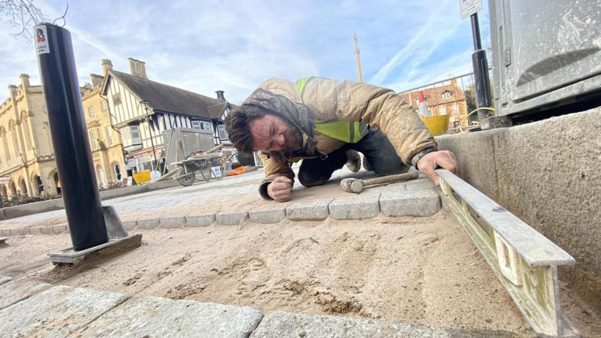 A photo taken from the ground of a men wearing a thick coat and a high-vis jacket placing a metal bar on to a pavement. A square with old buildings is behind him