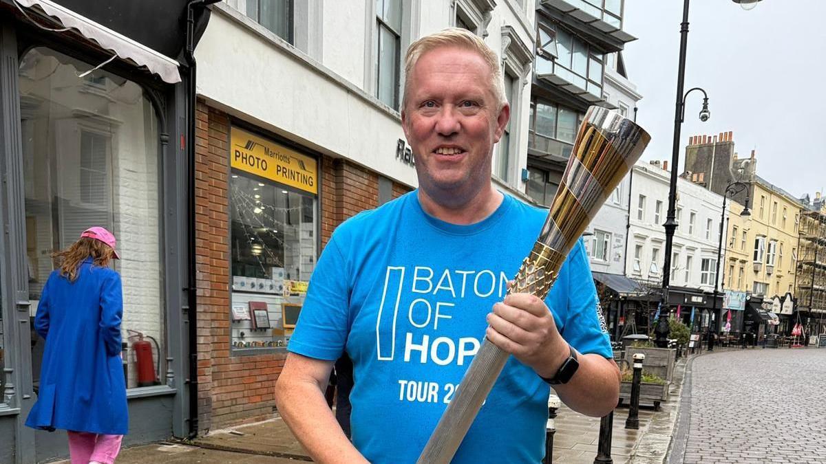 A man stands in a high street. He is wearing a blue t-shirt and holding a batton.