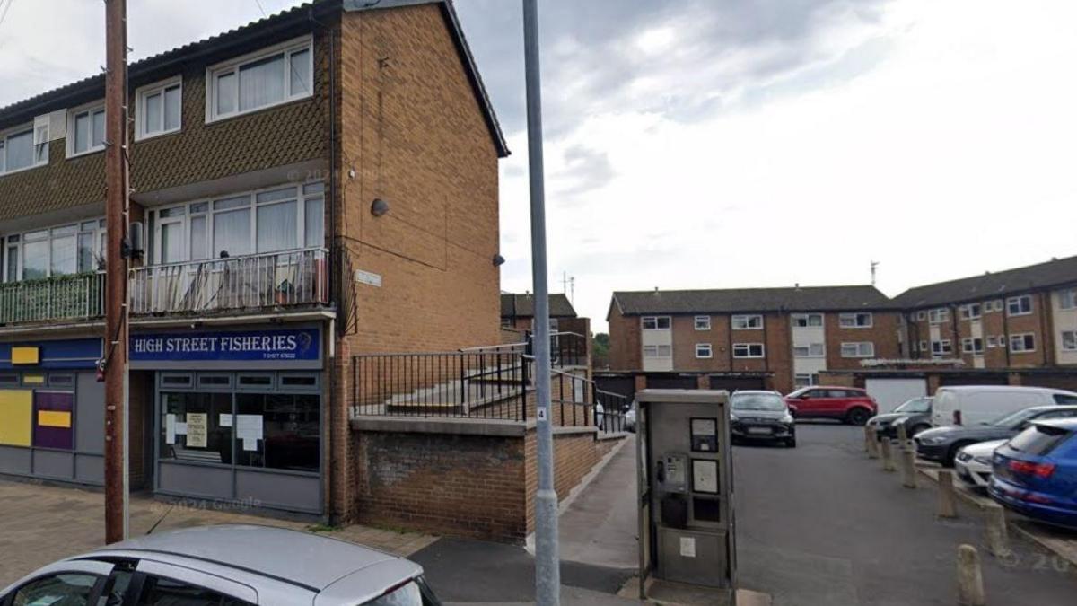 The location of the mural is a gable end of a brick building, in a residential area. There is a fish and chip shop in the building within a row of shops, and flats above. A phone box is in view as well as parked cars.