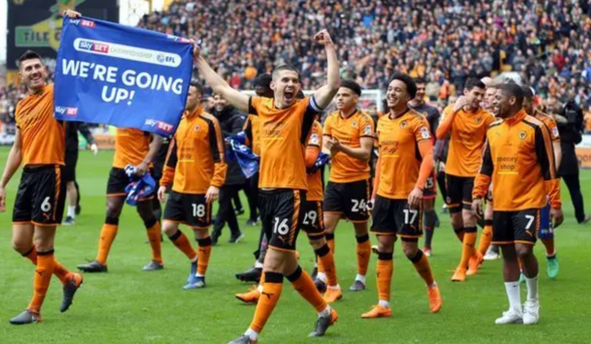 Wolves footballers, wearing old gold and black kits, on the pitch at Molineux on a lap of honour after winning promotion. Two of the players are holding a blue flag saying we're going up