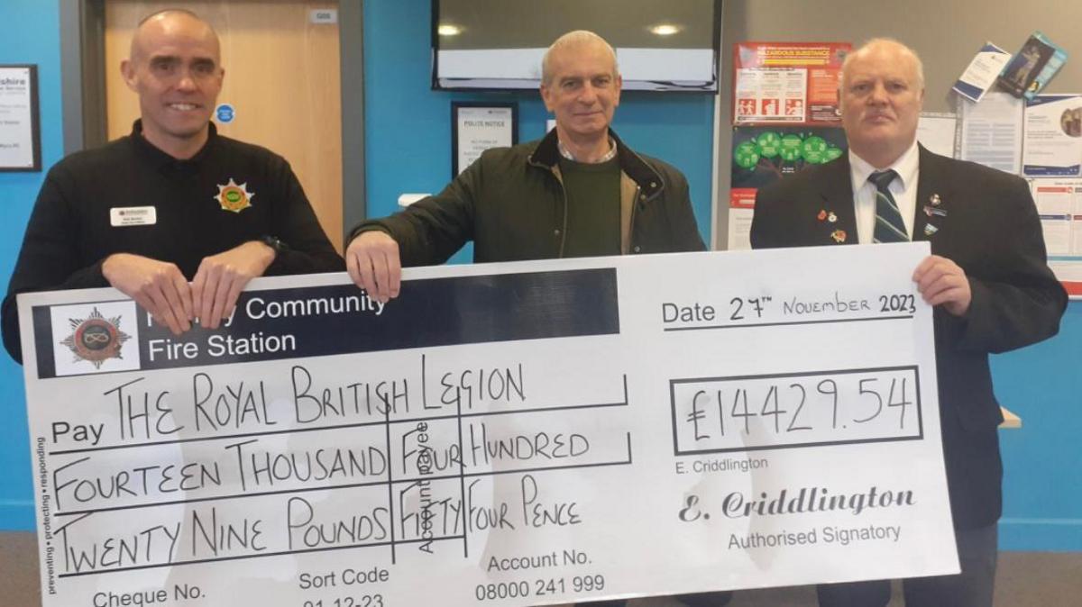 Three men stand in the reception room of a fire station holding a giant cheque. The cheque is made out to the Royal British Legion for £14,429.54. The man on the left is bald and wearing a jumper with a fire service emblem on his left breast and a name badge on the right. In the middle, a man has one hand on the cheque in a green wax jacket and green jumper. The man on the right is wearing a black suit, white shirt and a stripped tie.