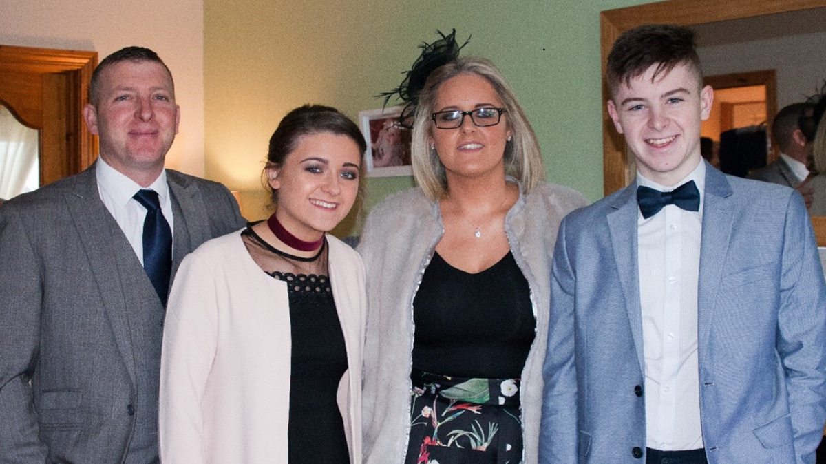 Neil Rooney stands in a grey suit with a navy tie on the left. On the right are his family. To his right are a brunette teenage girl, a blonde woman and a brown haired teenage boy. They are all wearing clothes for a special occassion. 