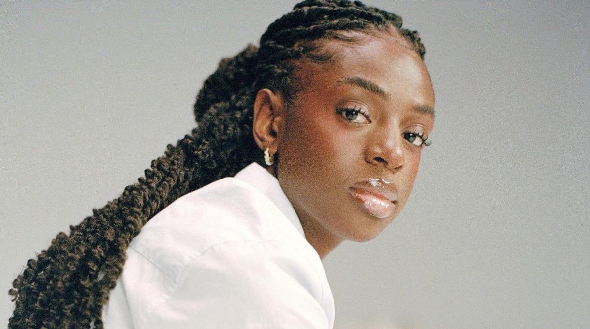 Cat Burns in a promotional shoot. She wears a white shirt and has her braided hair tied back. She looks at the camera with a serious expression over her right shoulder. She's pictured against a grey backdrop. 