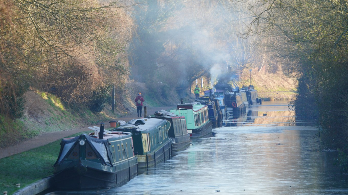 Several canal boats parked up in a line on the canal in a wintry scene with the water partially frozen over, the sunlight in the distance, and bare trees lining the bank