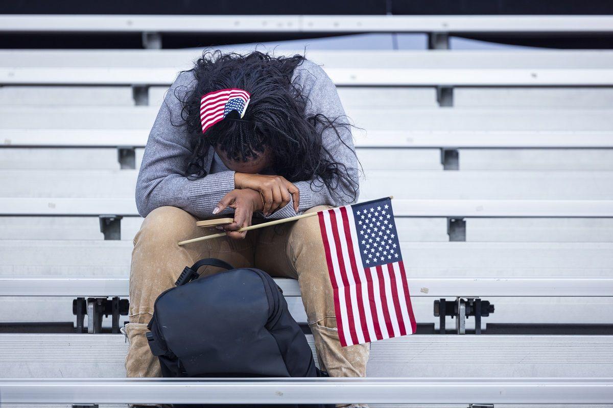 A woman holding an American flag with her head in her hands, sitting by herself in a row of bleachers