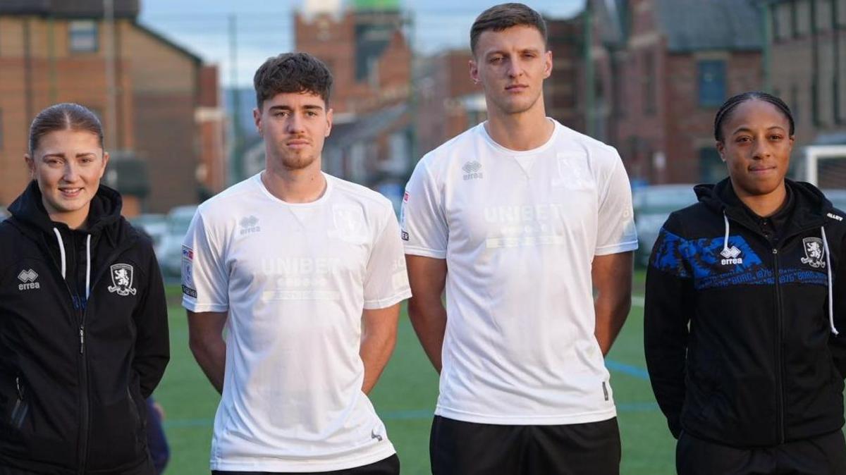 Middlesbrough players Hayden Hackney and Dael Fry wearing an all-white football shirt. They're standing between two women in black Middlesbrough tracksuits.