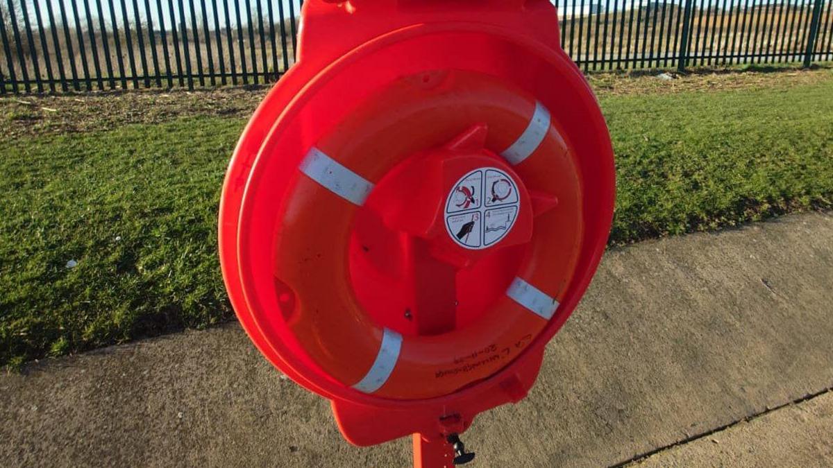 A circular red plastic cabinet on a pole, installed on a concrete path by a grass bank. The hinged lid is open and a red lifebuoy is visible inside, attached via white velcro.