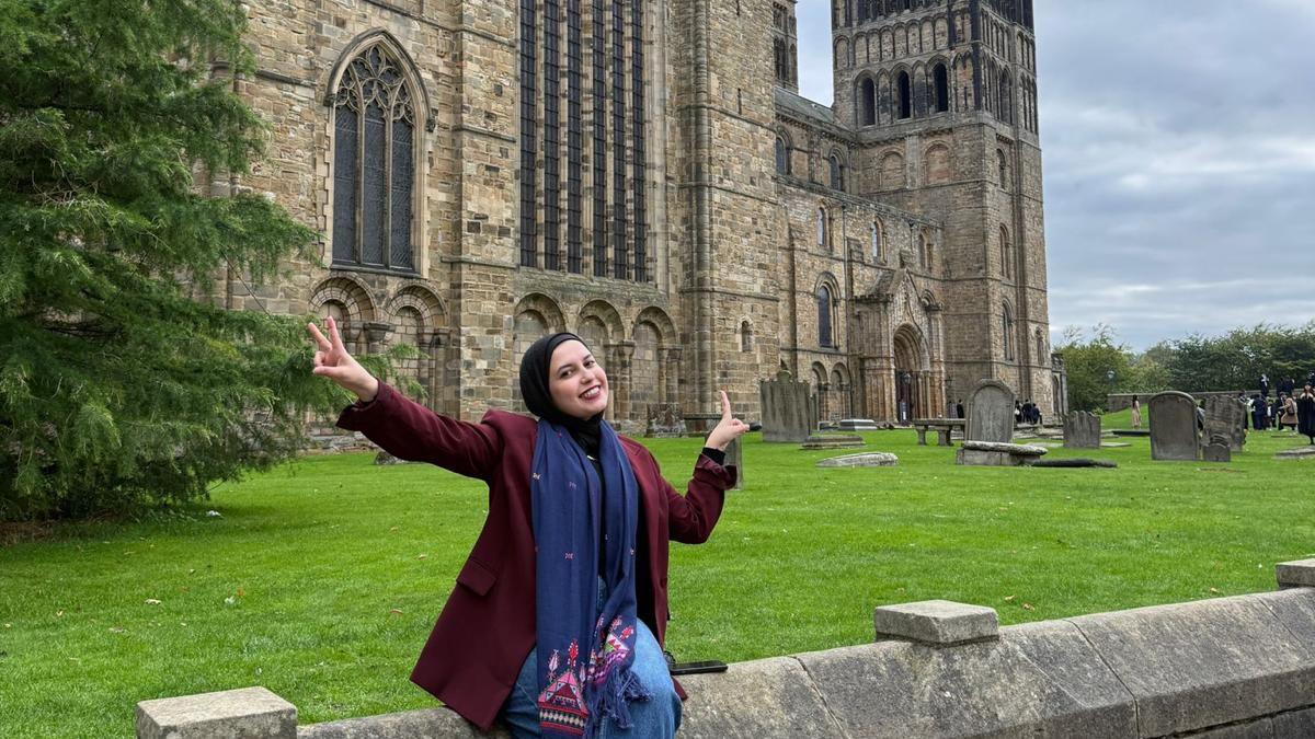 Sana el-Azab is sitting on a wall circling Durham Cathedral. She is smiling and is doing a peace sign with her hands. 