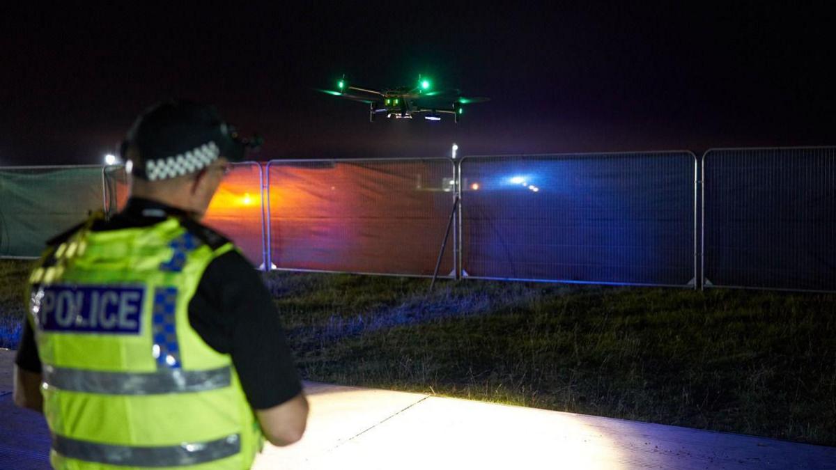 Image shows a police officer in a high-vis vest and headtorch operating a drone at night over a field.