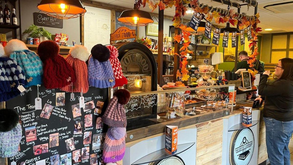 A woman wearing jeans, a dark jumper and glasses is standing at the counter of the coffee shop. A man can be seen preparing an order. There are cakes behind glass on the counter, along with a hot food cabinet, with signs for bacon sandwiches. There is also a board with photos of some of the dog visitors to the cafe.