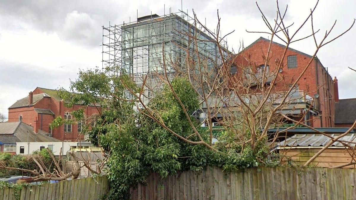 A view of the rear of Jacob's Court in Hereford. Scaffolding covers the tower block and other warehouse buildings are nearby. There is a fence in front of the site, with a tree growing over it.