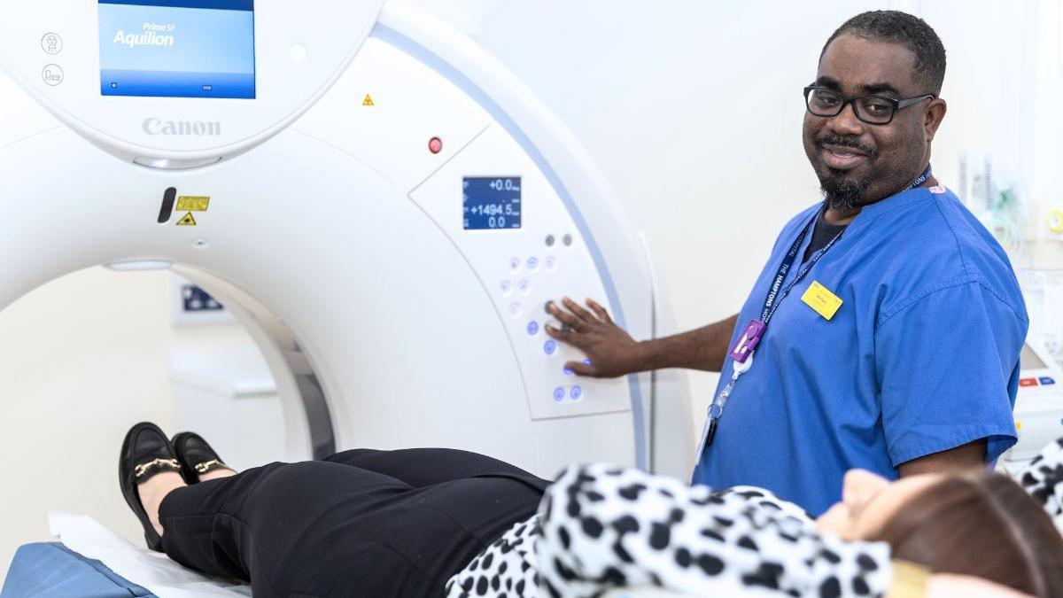 A man in blue scrubs is helping a patient, who is lying on their back, go into an MRI scanner.