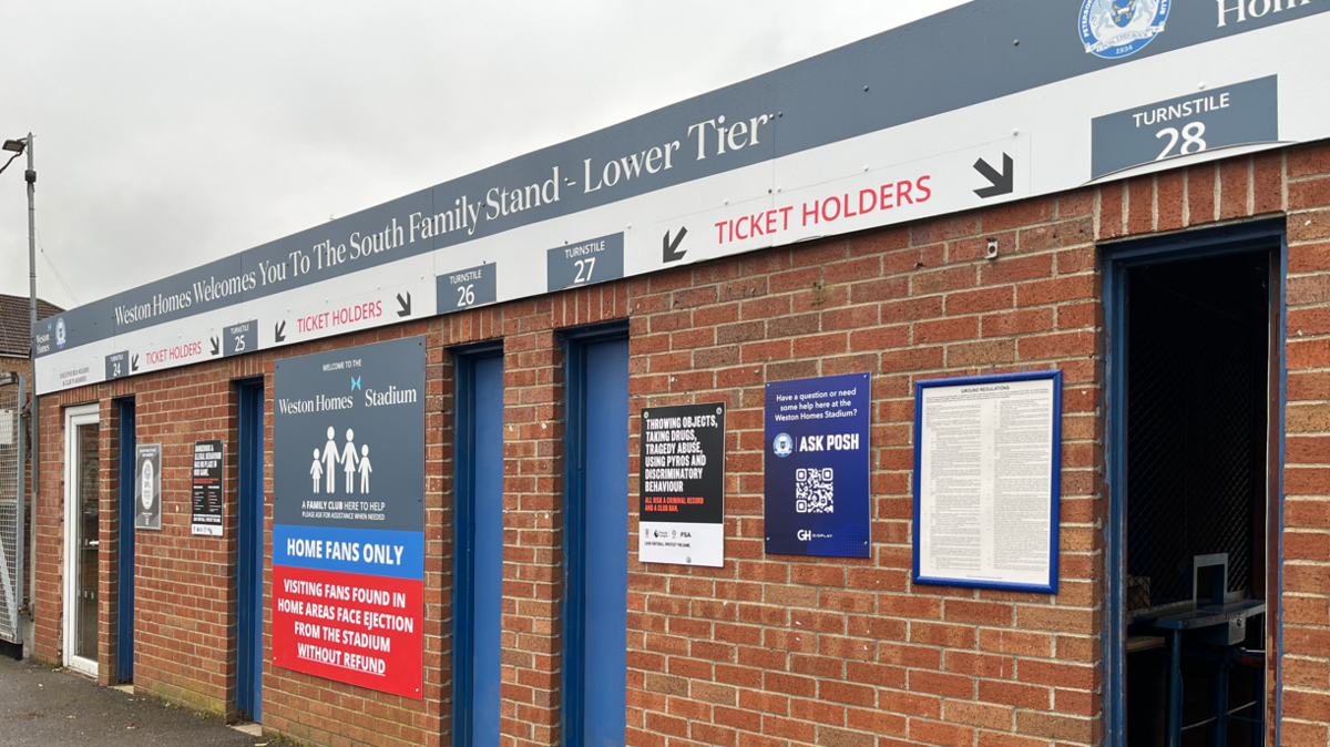 Outdoor view of the turnstile building. It is red bricked with a sign over the door directing ticket-holders though the turnstiles. There is a sign on the wall reading 'Home fans only'. There are five thin blue doors for fans to push through to enter the stadium. 