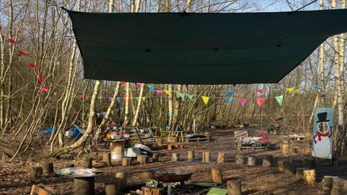 A small camp in the middle of a forest with wooden tree trunk seats and a firepit sitting underneath a green tarpaulin shelter.