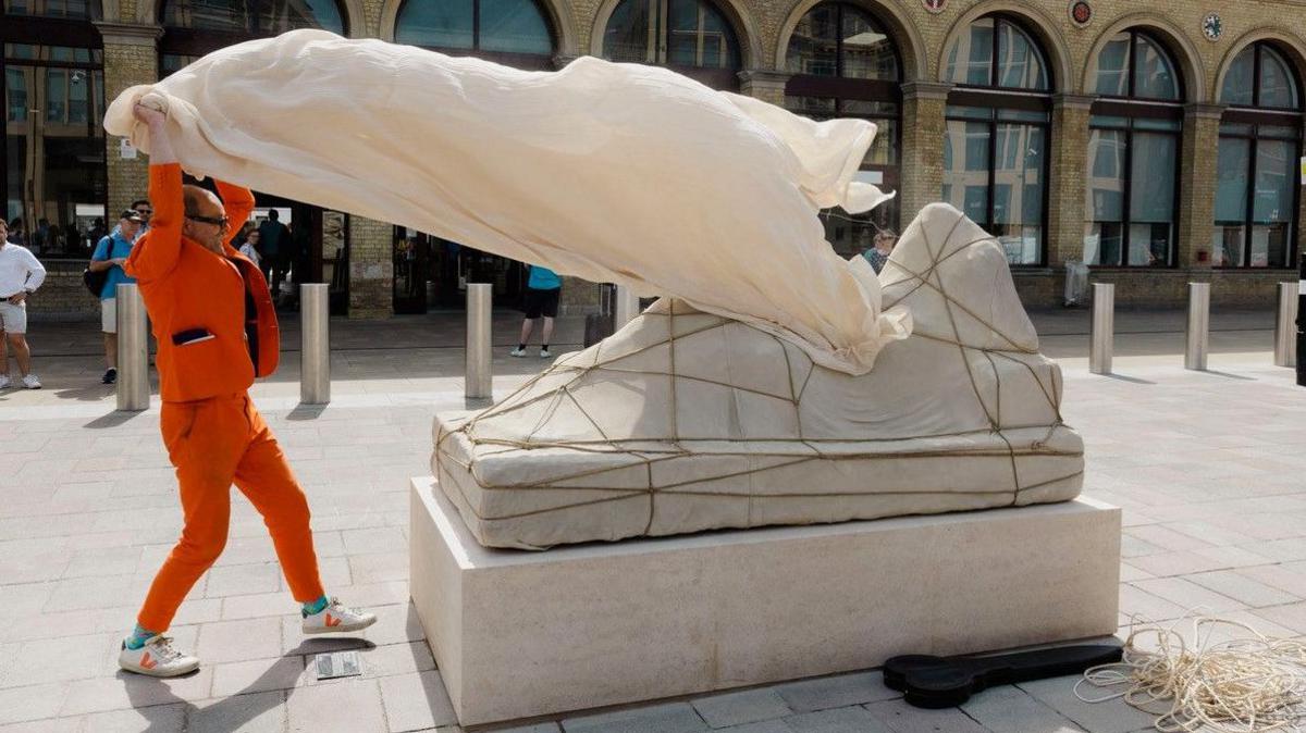 Gavin Turk, who is wearing an orange suit, pulls a dust sheet off the Ariadne sculpture. The sculpture is made of grey stone and laced on top of a large rectangle block. The sculpture looks like a woman who has been covered with a sheet and rope. In the background is a brick building with lots of windows.