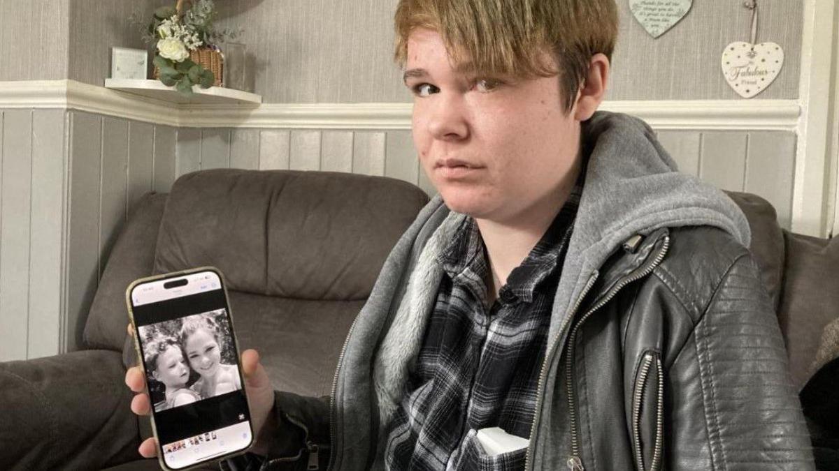 A woman on a sofa is looking at the camera and holding a mobile phone, which has a photo on it.