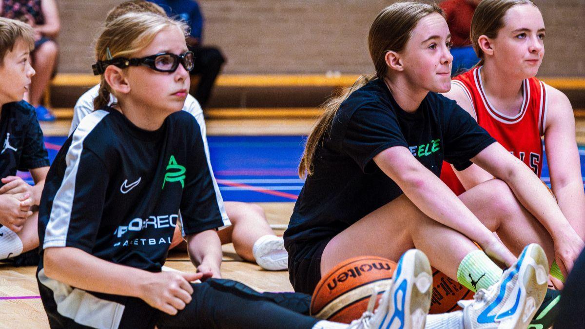 Three girls, one with long blonde hair and two with long dark hair, sit on a basketball court with an orange ball between them. The one nearest the camera is wearing blue and white trainers.