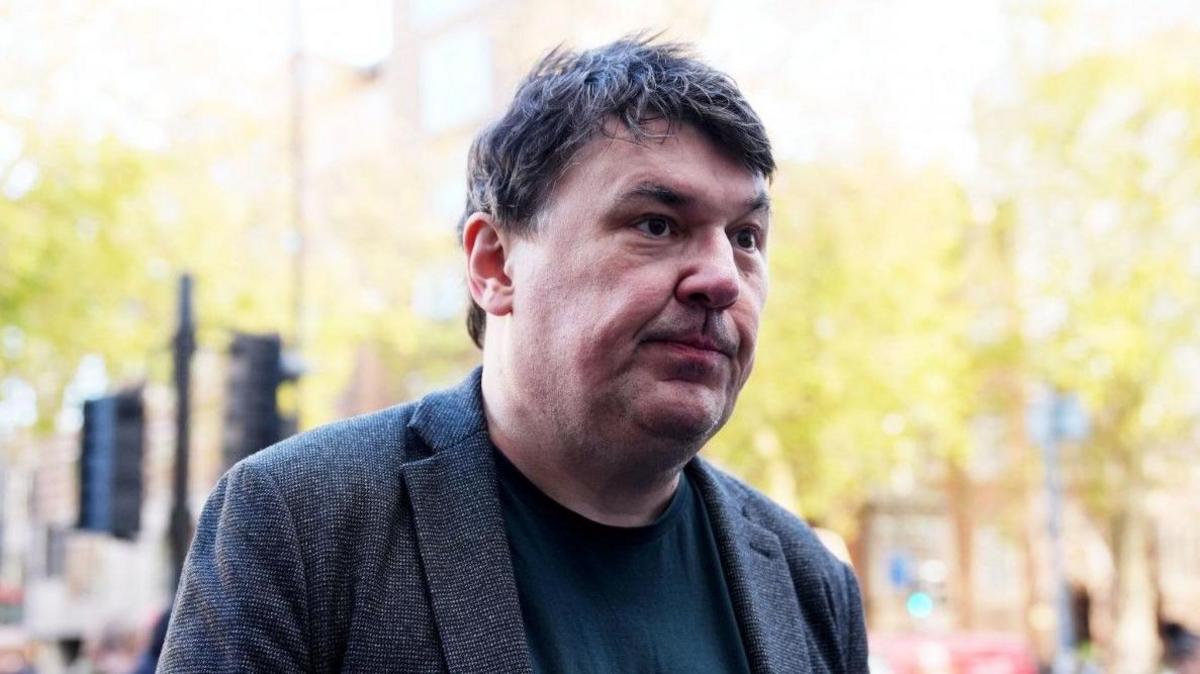 Graham Linehan, a man with dark hair, wearing a grey jacket and dark top.