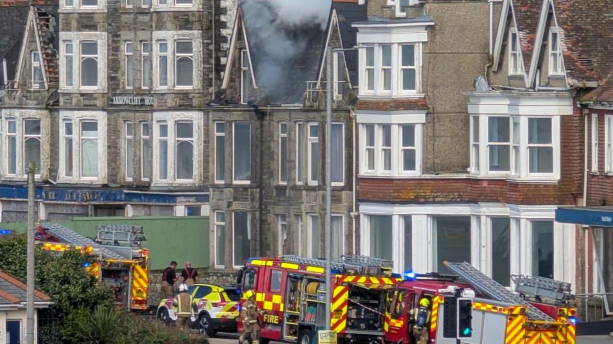 Three fire engines and other emergency vehicles can be seen in front of a three storey building. There is smoke coming out of the roof.