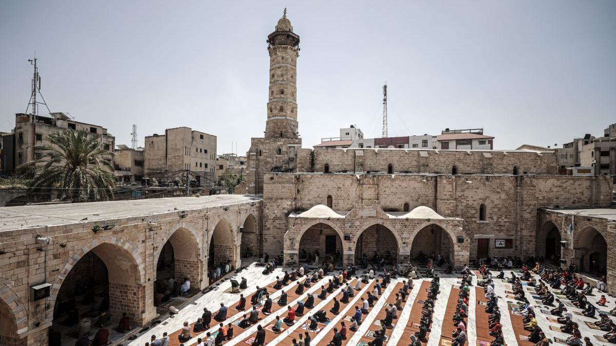 A stone coloured mosque in the shape of a square, there are arches surrounding the square. In the centre of the mosque is rows of people praying on mats. In the left hand corner of the mosque is a stone coloured tower, the background is also filled with various stone buildings.