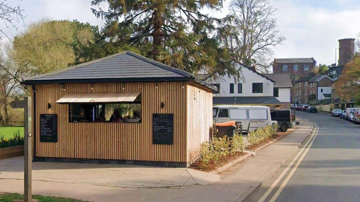 A wooden cafe kiosk next to a road. It has light wood walls and a black pointed tiled roof. There is a large window propped open and two blackboards on the outside with white chalk writing on them. The road, on the right hand side, has cars parked on its right side, with houses in the background.