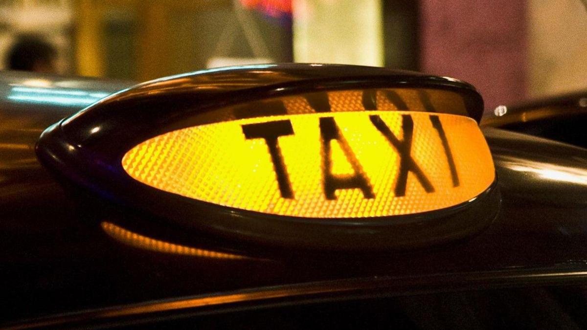 A close-up photo of a lit-up yellow taxi sign on the top of a vehicle. It says taxi in capital letters in the foreground, while the background is blurred.
