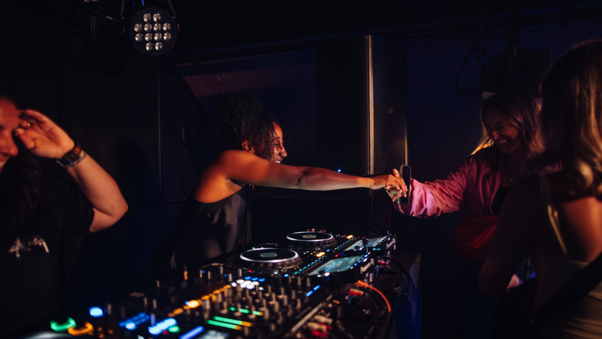 Jamz Supernova is pictured standing over decks interacting with fans, smiling broadly and leaning over to shake hands with a fan.