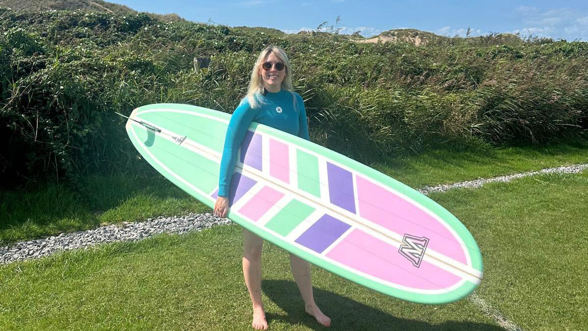 Gwenno Haf Hughes smiles at the camera with her new surfboard. She is standing on grass. The surfboard is turquoise, pink, purple and white and has a W on the top. Gwenno is blonde and is wearing a turquoise wetsuit. 