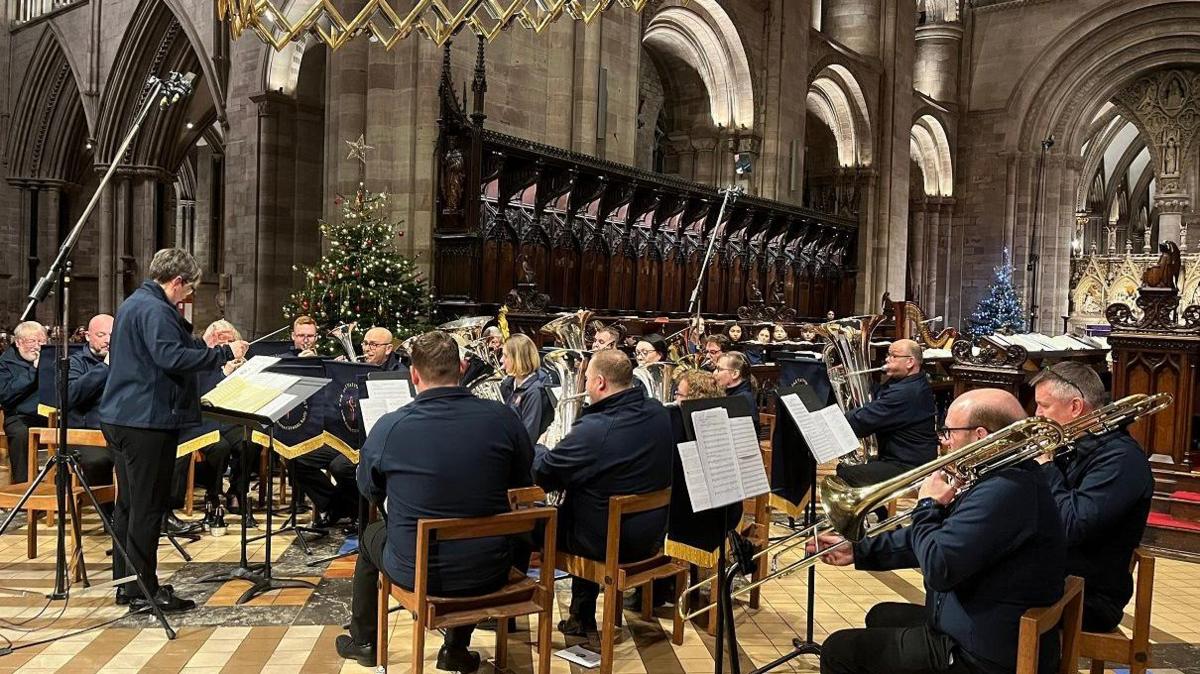 Musicians sit on wooden chairs playing instruments with sheet music on stands before them. Their instruments include tubas and trombones and they are conducted by a person with short dark hair with a microphone above them.