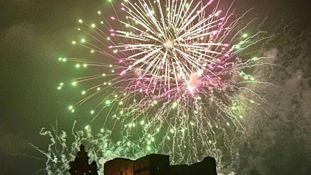 A big burst of a pink firework in the sky, sending out streams of light, with a slightly bigger but fainter green firework burst just behind it. They appear to be above some historic building, maybe a castle, with stone walls visible at the bottom.