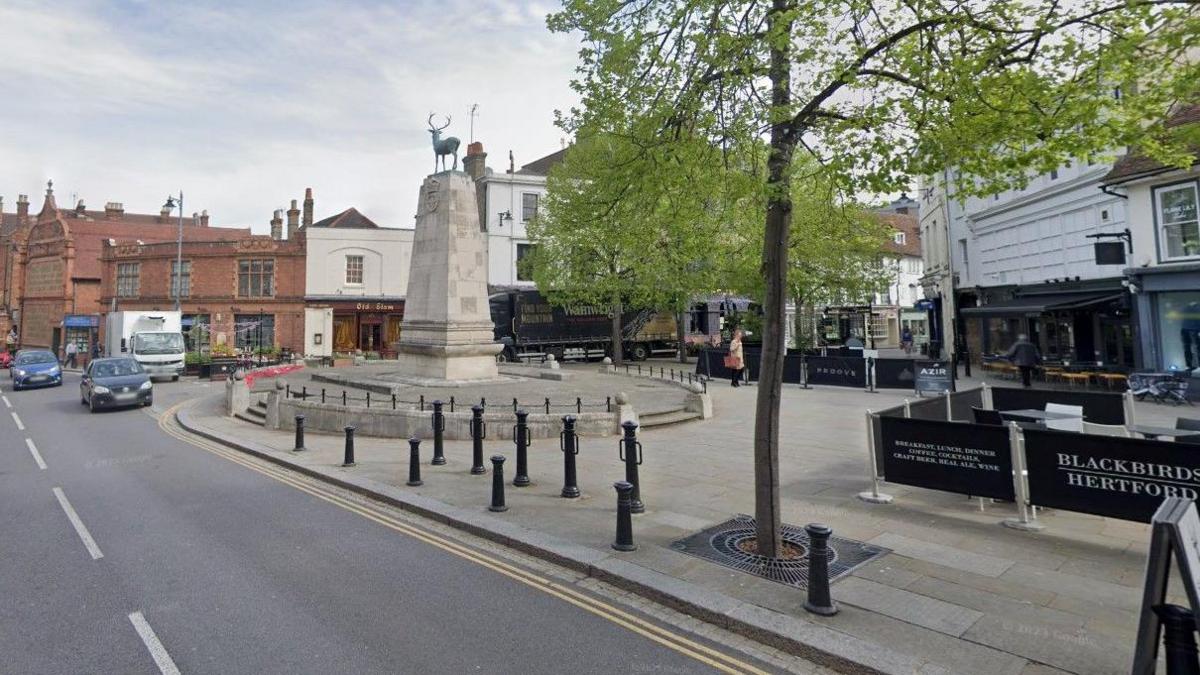 Parliament Square in Hertford, with three vehicles in the distance, a war memorial and a tree