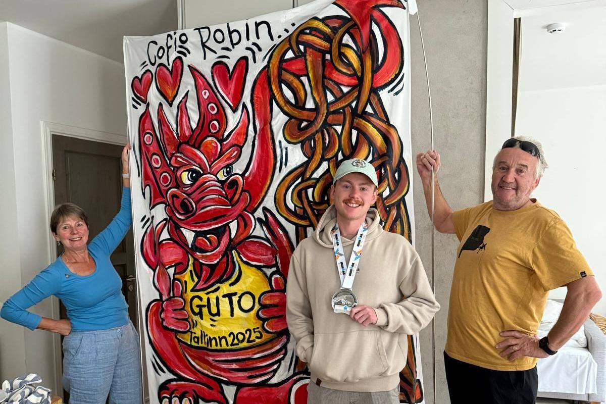Llun o rieni Guto'n dal baner mawr y tu ol i Guto sy'n dal medal Ironman