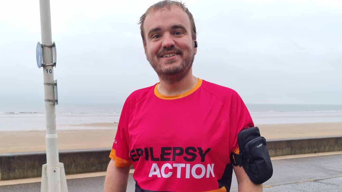 A man wearing a pink sports top smiles for the camera next to the seaside.