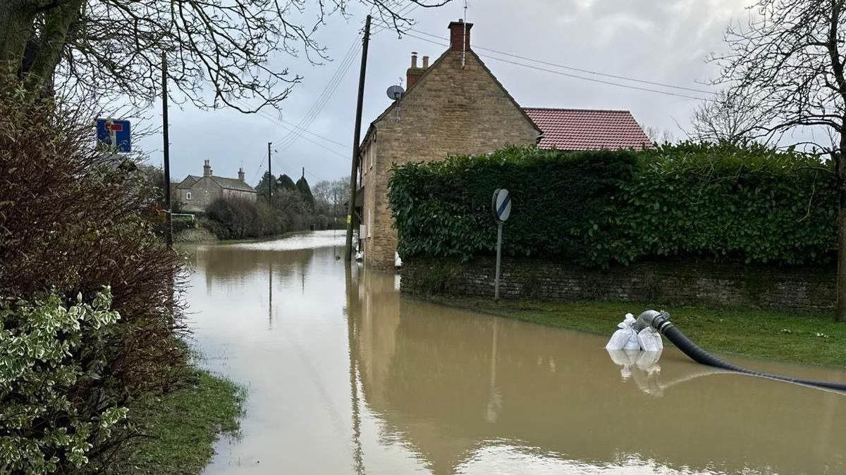 Brown flood water covers a road in Tallington, Lincolnshire. To the right is a tall hedge and in the centre a house with sand-coloured bricks