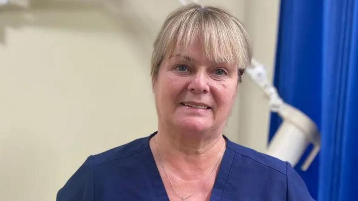 A woman, with blonde hair, wearing dark blue scrubs.