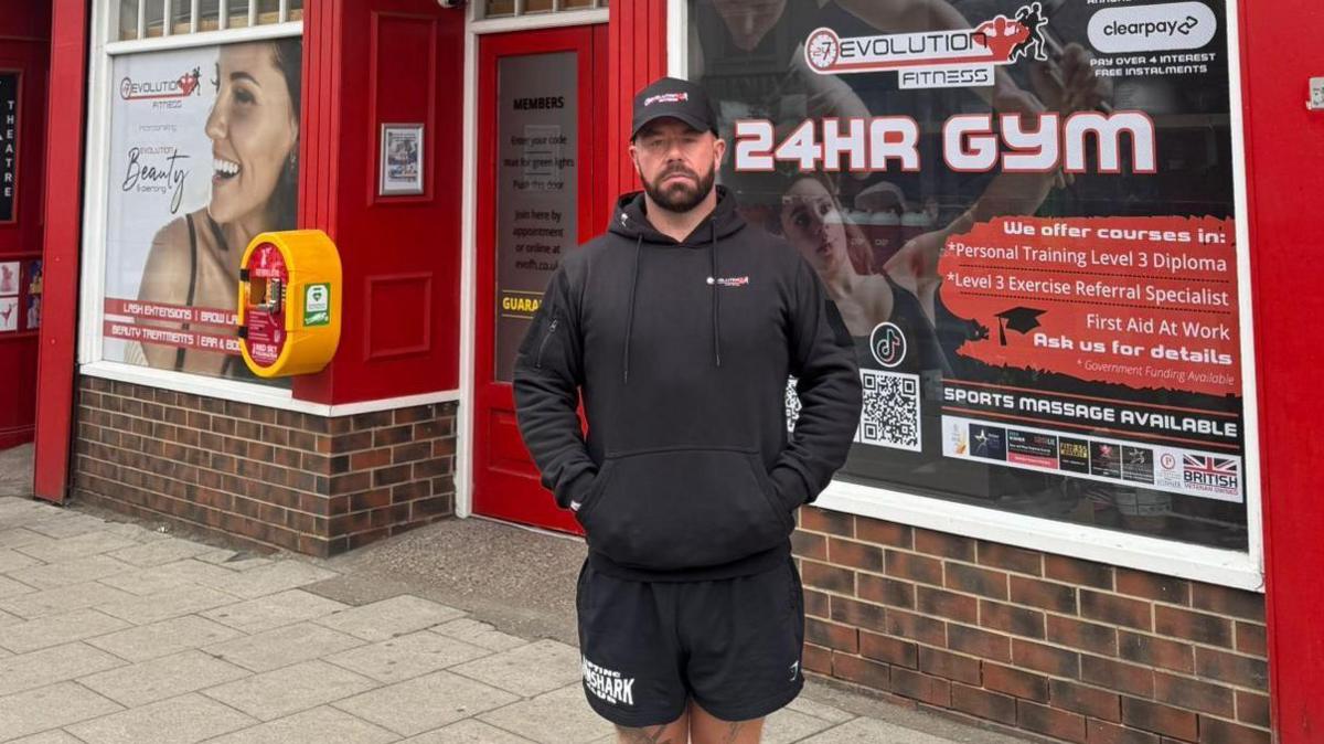 Micky Donkin is standing in front to the Evolution Fitness Gym which has a red painted exterior with various stickers in the window, including one that says it is a 24-hour gym. Mr Donkin has a black beard and is wearing a black cap, hoody and shorts.