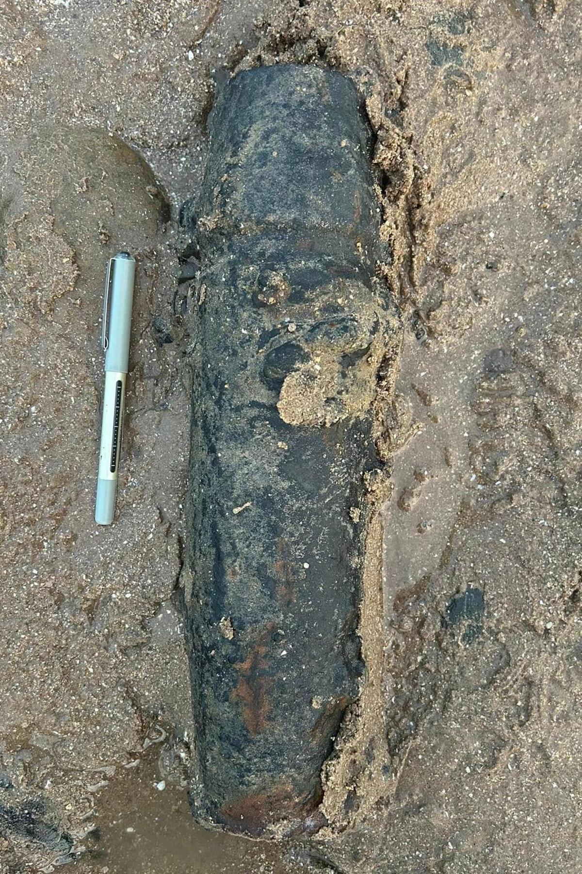 The shell, which is black and battered, partly covered in the sand on the beach. It is next to a pen which is about a third of its height.