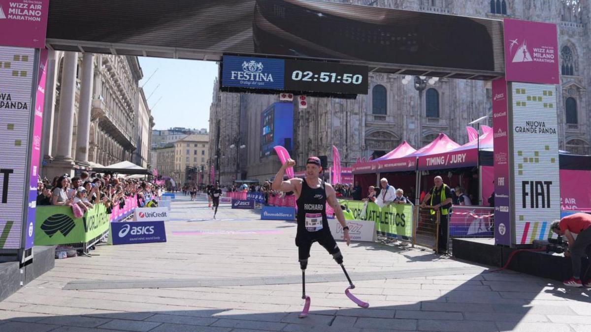 Richard Whitehead, he is at the marathon finish line in a city, you can see his time above his head which is 02:51:50, he wears a vest, cap and has running blades.