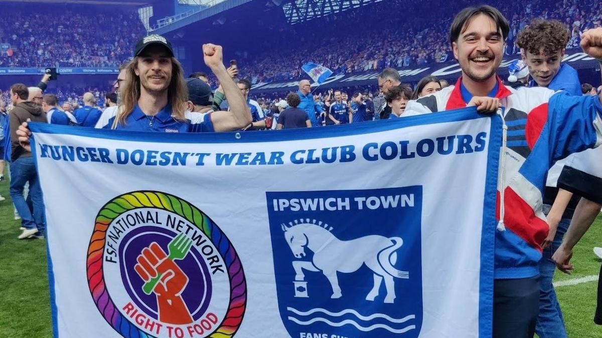 Charlie Nixon holds the hunger doesn't wear club colours flag along with another fan. They stand on the Portman Road pitch and wave their spare hands in the air in joy. 